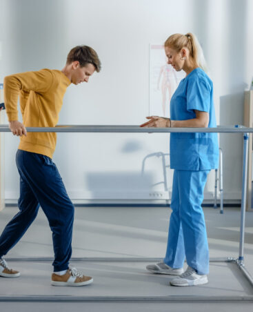 Hospital Physical Therapy: Strong Patient with Injury Making First Steps, Walks Holding for Parallel Bars. Physiotherapist, Rehabilitation Doctor Assist, Help Disabled Person