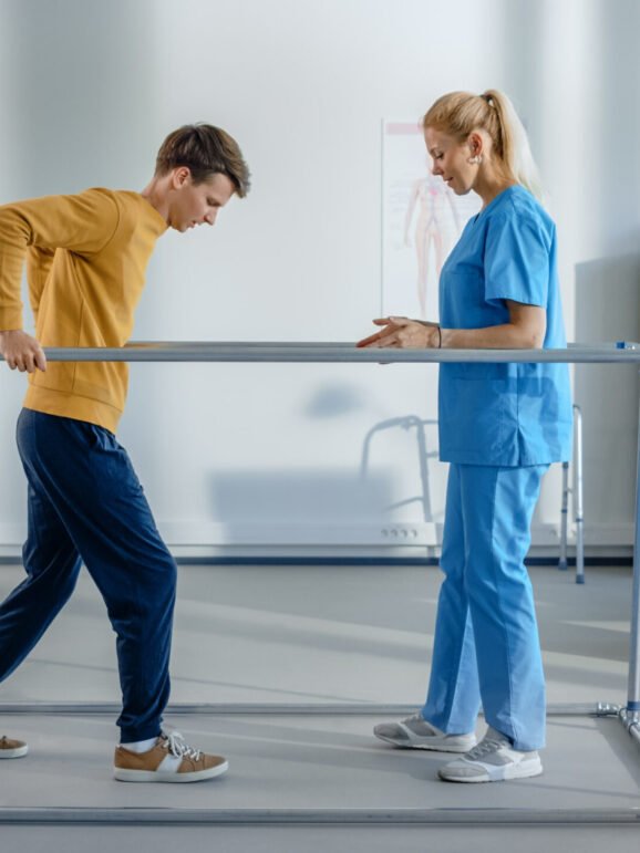 Hospital Physical Therapy: Strong Patient with Injury Making First Steps, Walks Holding for Parallel Bars. Physiotherapist, Rehabilitation Doctor Assist, Help Disabled Person