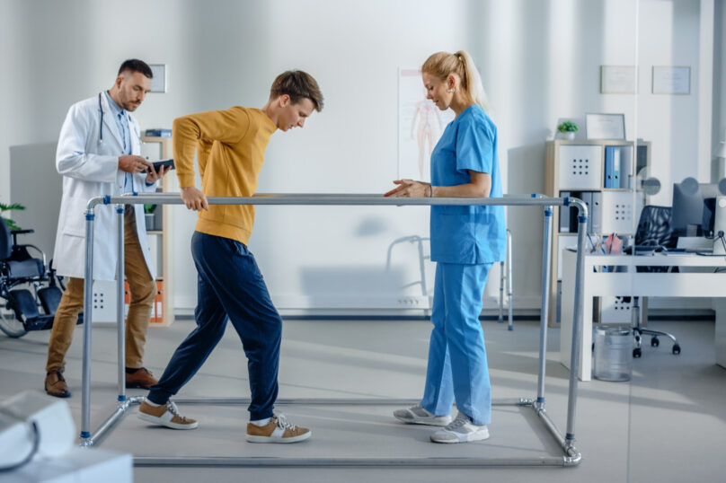 Hospital Physical Therapy: Strong Patient with Injury Making First Steps, Walks Holding for Parallel Bars. Physiotherapist, Rehabilitation Doctor Assist, Help Disabled Person