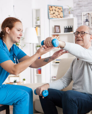 Senior man in nursing home with doing physical therapy with help from nurse using dumbbells.